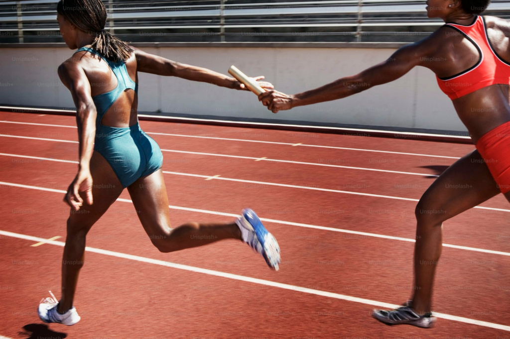 Two runners are on the track, with one handing a baton to the other in front of her