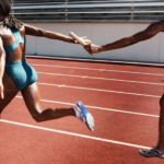 Two runners are on the track, with one handing a baton to the other in front of her