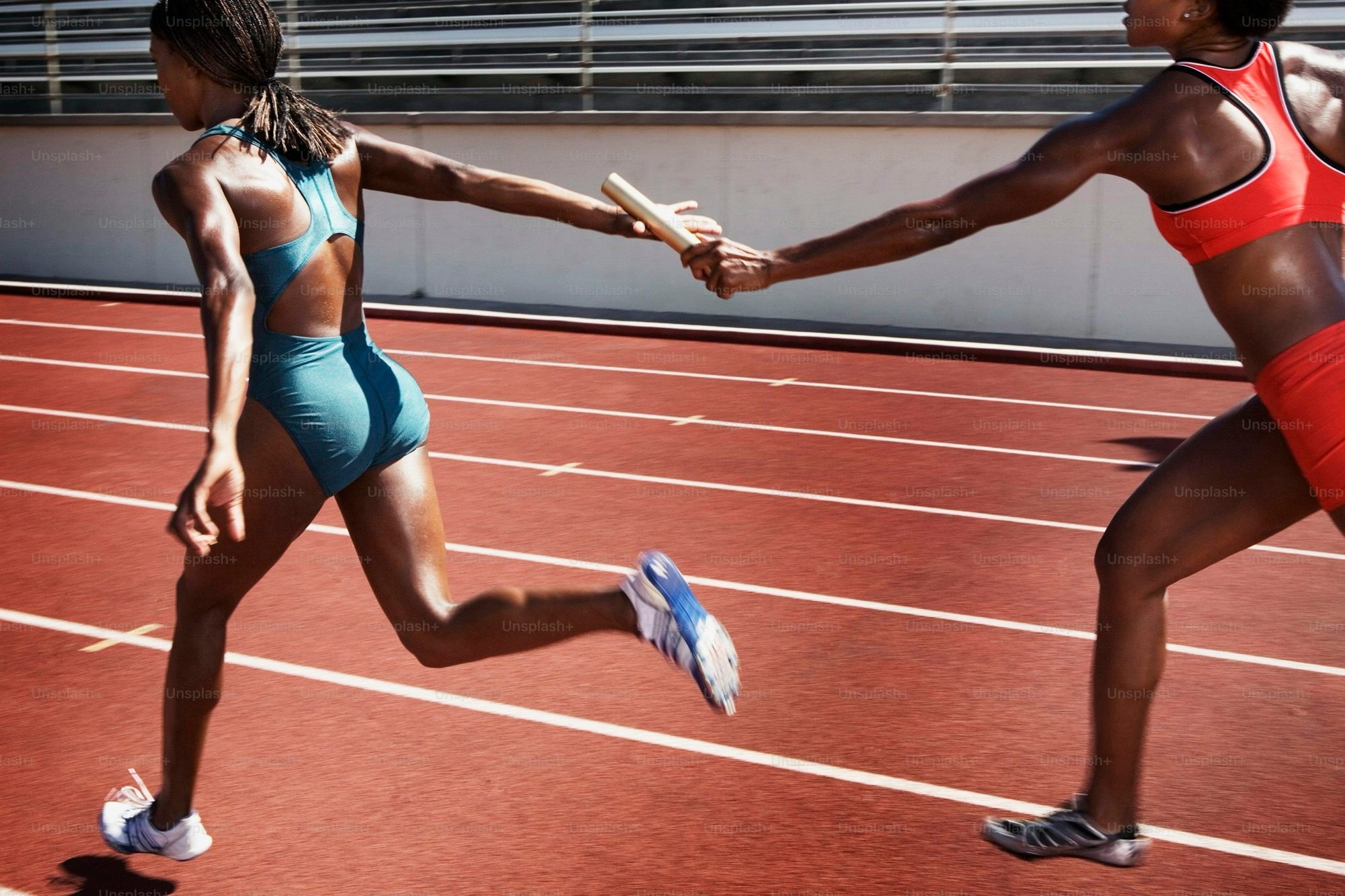 Two runners are on the track, with one handing a baton to the other in front of her