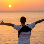 A man stands at the edge of some water, with the sun just above the horizon, holding his arms out wide