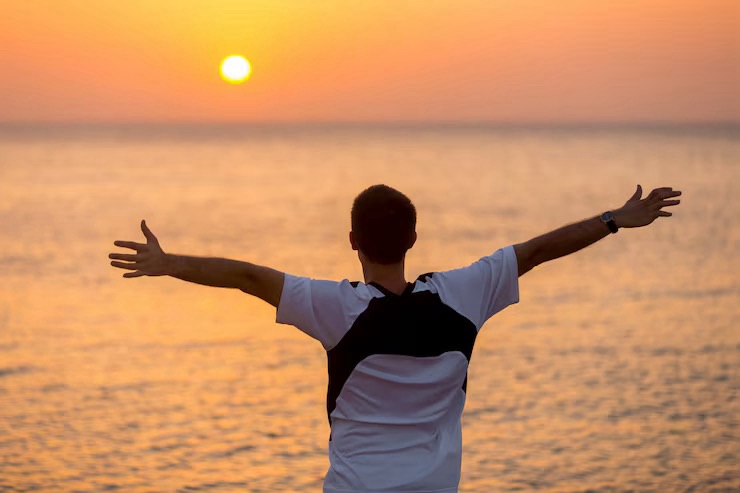 A man stands at the edge of some water, with the sun just above the horizon, holding his arms out wide